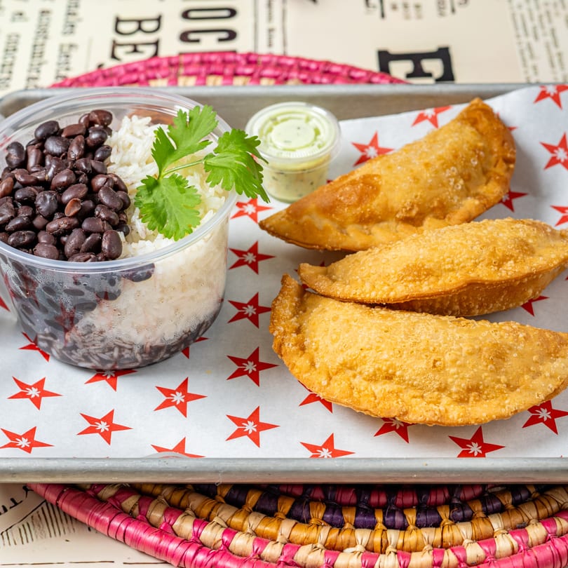 Cuban black beans and rice in a clear container with three golden empanadas and a dipping sauce.