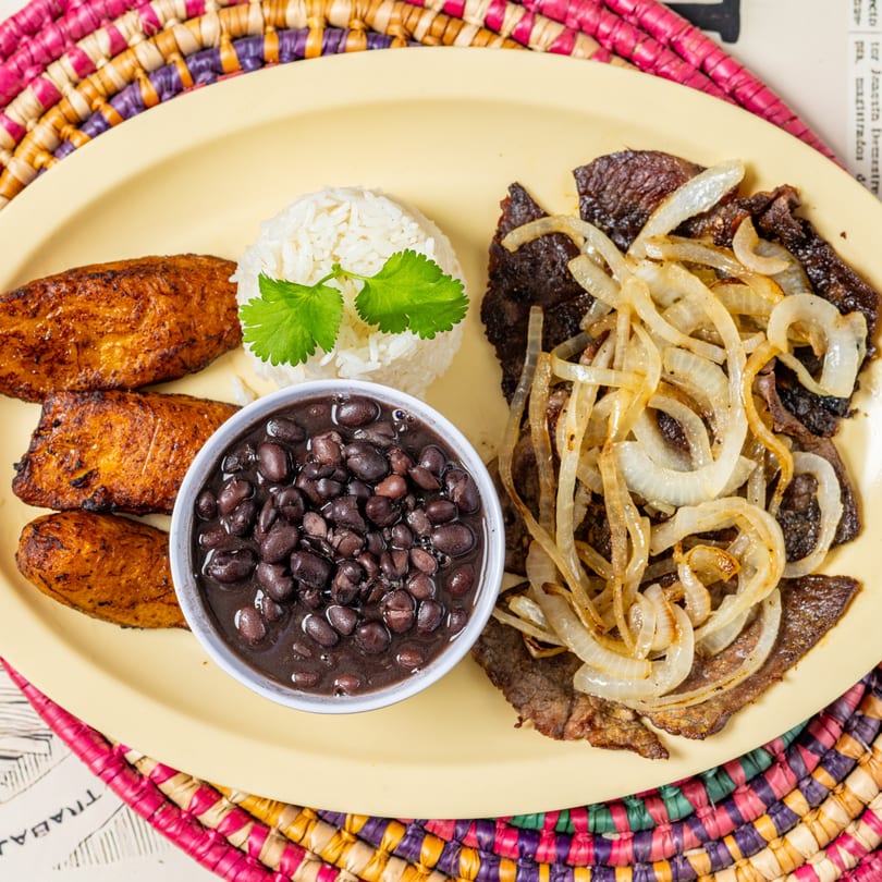Cuban steak with onions, rice, black beans, and fried plantains on a yellow plate.