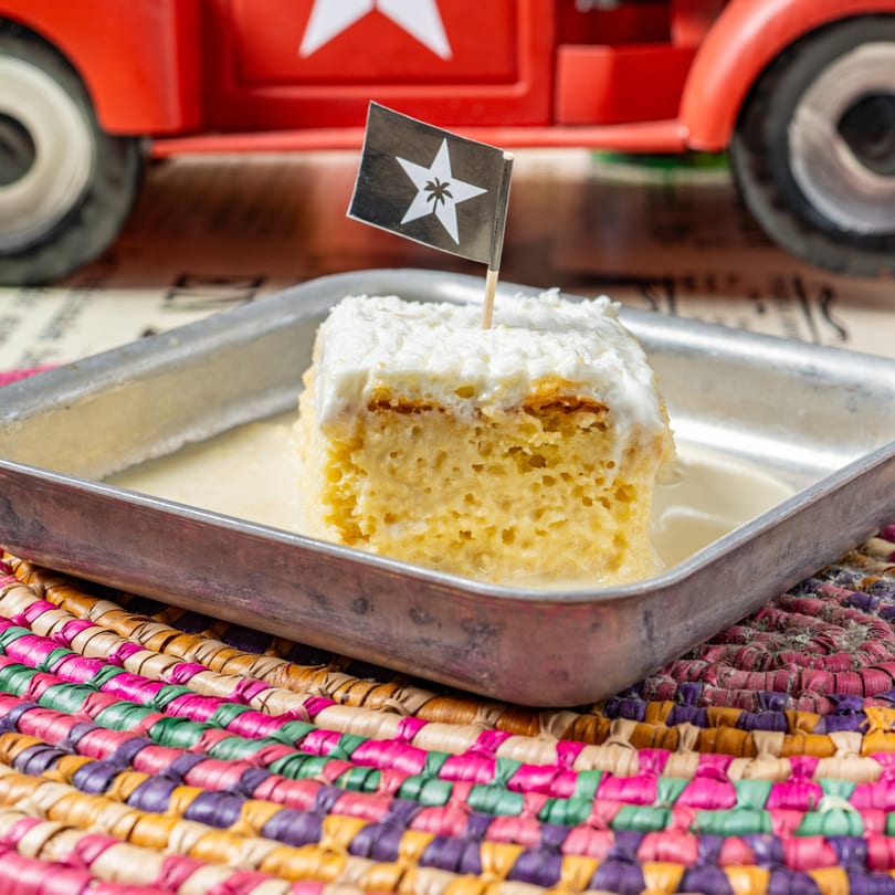 A slice of tres leches cake with a Cuban flag pick, served in a metal tray.